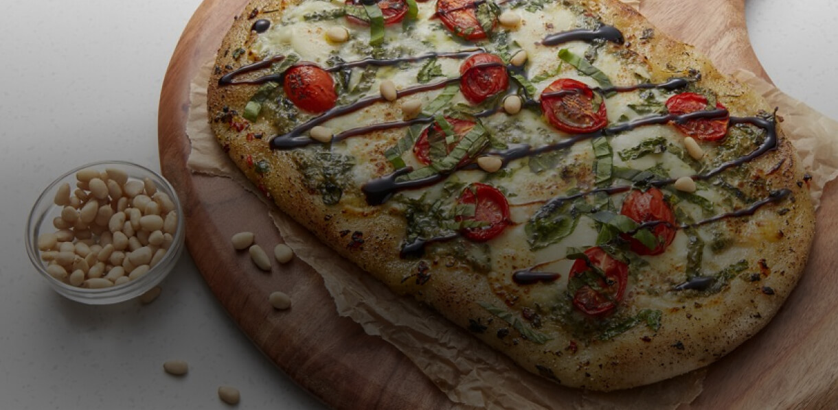 A flatbread pizza on a wood serving board next to a bowl of pine nuts. 