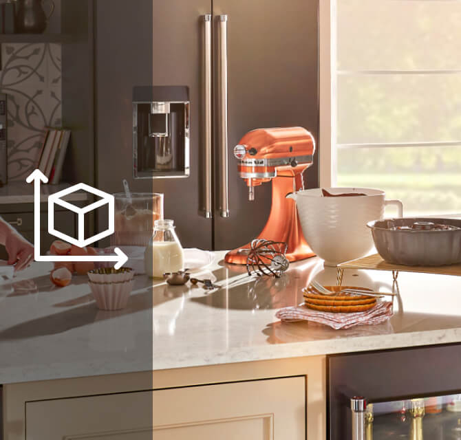 A person drizzling red icing on a cake and baking ingredients and a KitchenAid® stand mixer on the counter