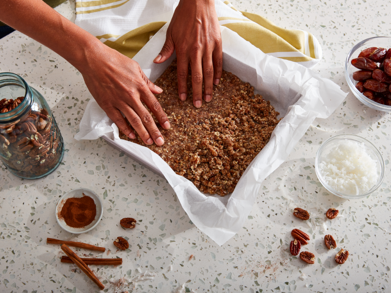 A pair of hands putting a baking dish together with ingredients consisting of coconut, dates, pecans and cinnamon