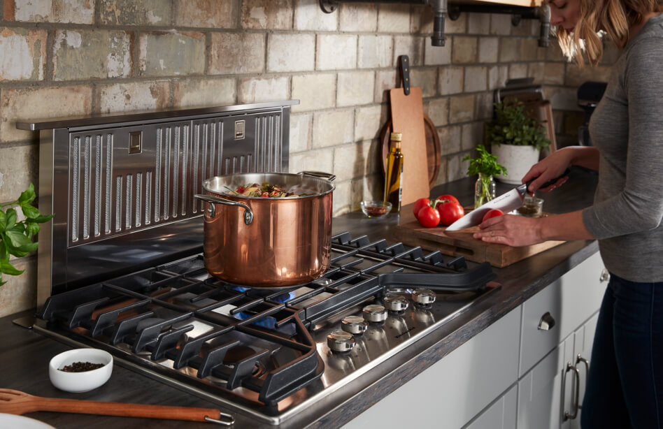 Bronze pot resting on center grate of a cooktop and person slicing tomatoes on a cutting board