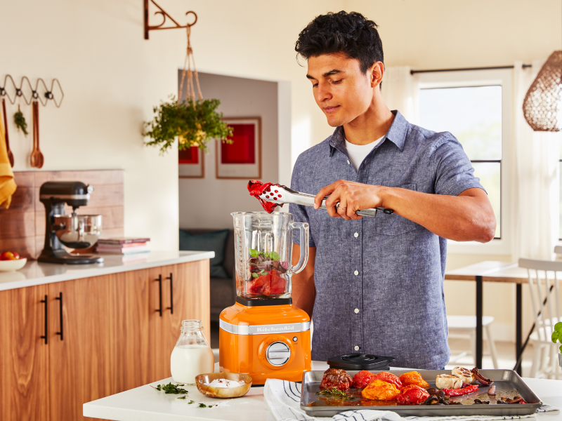 A man adding roasted vegetables to a KitchenAid® blender in a modern kitchen.
