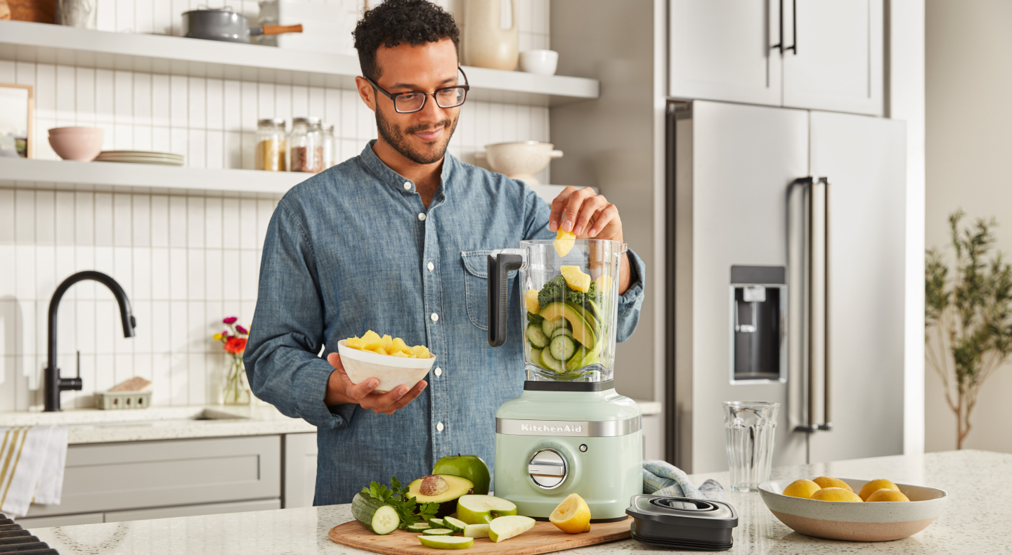 A man putting fresh vegetables into a KitchenAid® blender in a modern kitchen.