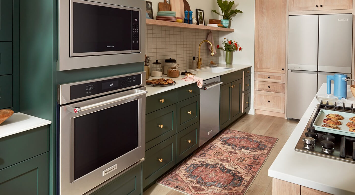 A galley kitchen with forest green cabinetry, white countertops, stainless steel appliances and white tile backsplash on one side, and a kitchen island with white countertops and a stove top on the other