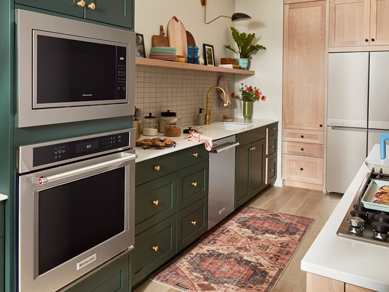 A galley kitchen with forest green cabinetry, white countertops, stainless steel appliances and white tile backsplash on one side, and a kitchen island with white countertops and a stove top on the other