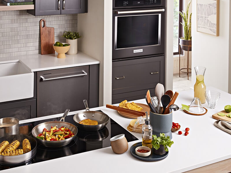 A galley kitchen with dark brown cabinetry, white countertops, a sink and built-in oven on one side, and an island with white countertops and a stovetop on the other