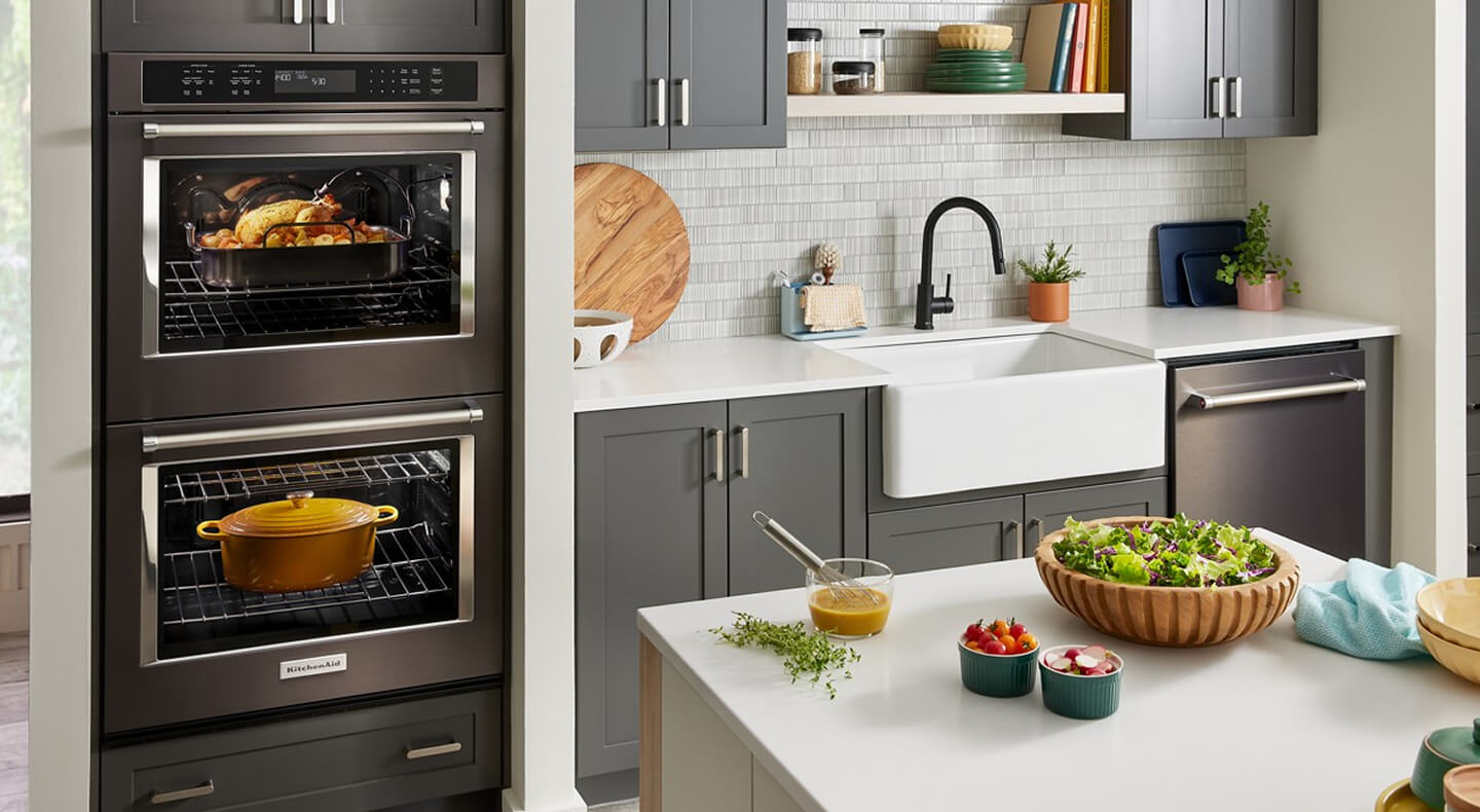 A kitchen with a double oven, apron sink, gray cabinets and a dishwasher against one wall, and an island with a white countertop across the way