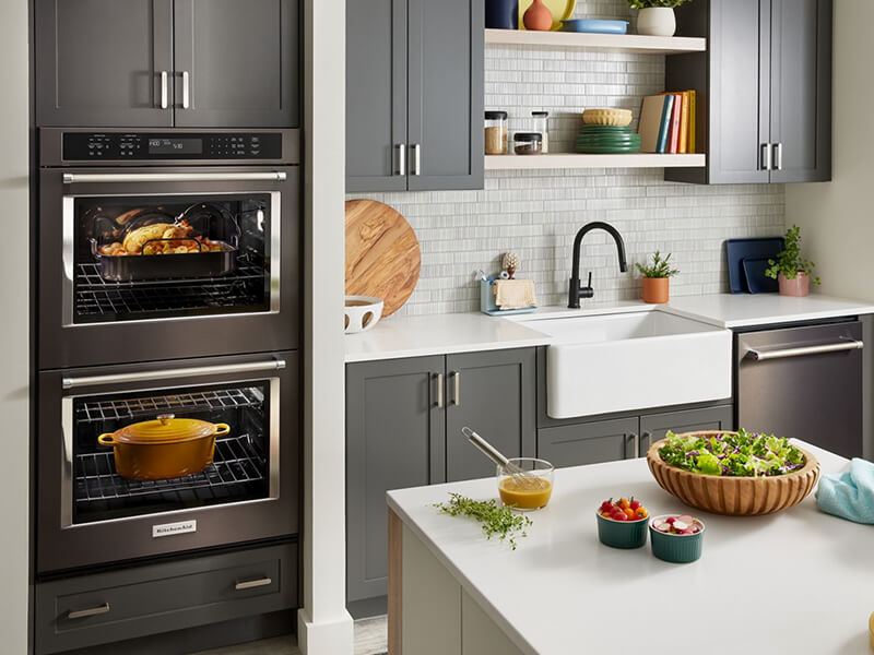 A kitchen with a double oven, apron sink, gray cabinets and a dishwasher against one wall, and an island with a white countertop across the way
