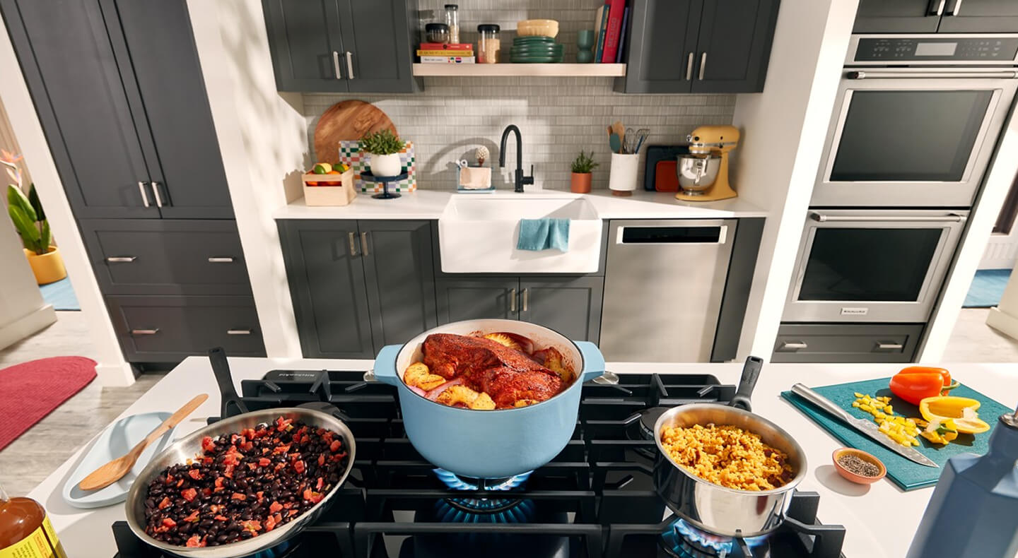 A galley kitchen with dark gray cabinets, dishwasher and double oven on one side, and an island with a stovetop loaded with full pots and pans on the other