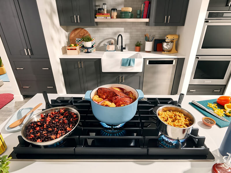 A galley kitchen with dark gray cabinets, dishwasher and double oven on one side, and an island with a stovetop loaded with full pots and pans on the other