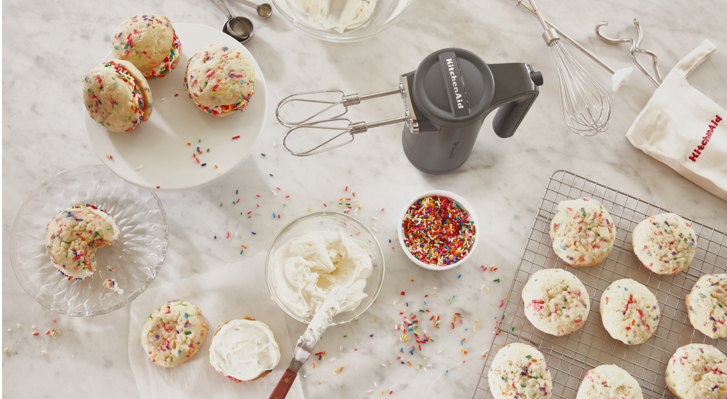 Cordless KitchenAid® hand mixer next to creamy frosting and fresh baked sandwich cookies with colorful sprinkles.