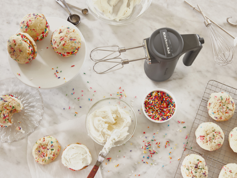 Cordless KitchenAid® hand mixer next to creamy frosting and fresh baked sandwich cookies with colorful sprinkles.