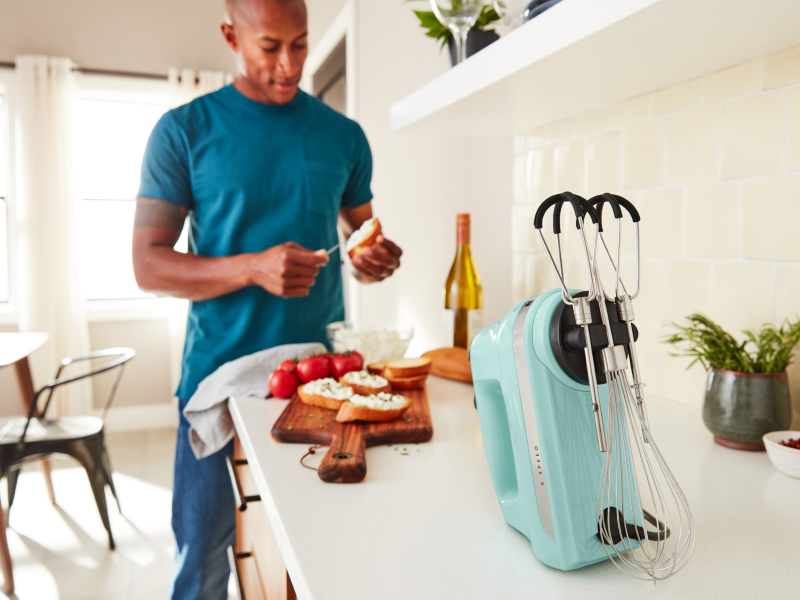 A man in a modern kitchen spreading goat cheese on a crostini next to a KitchenAid® hand mixer. 