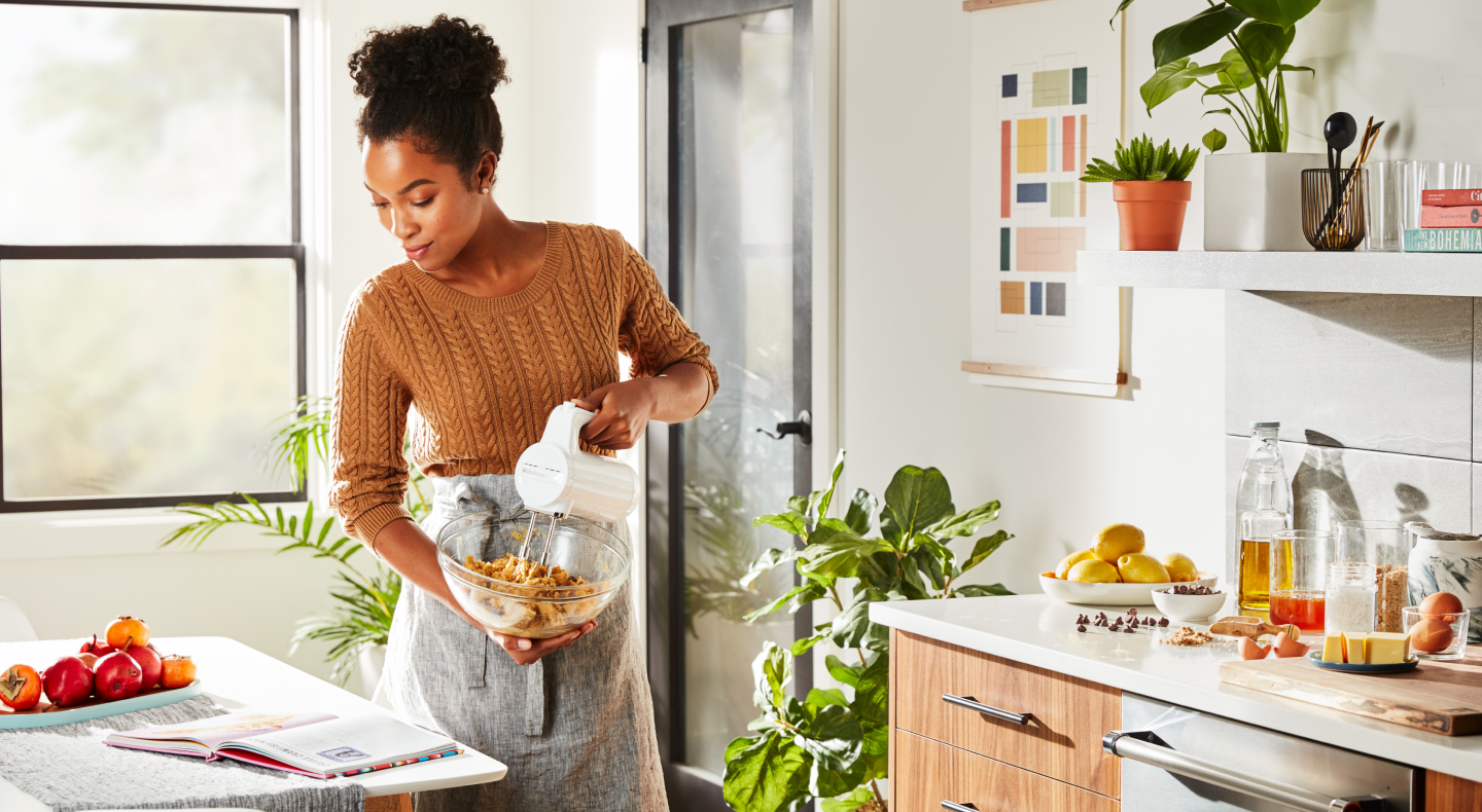 A woman in a modern kitchen reading a cookbook and holding a mixing bowl while using a Cordless KitchenAid® hand mixer to mix cookie dough.