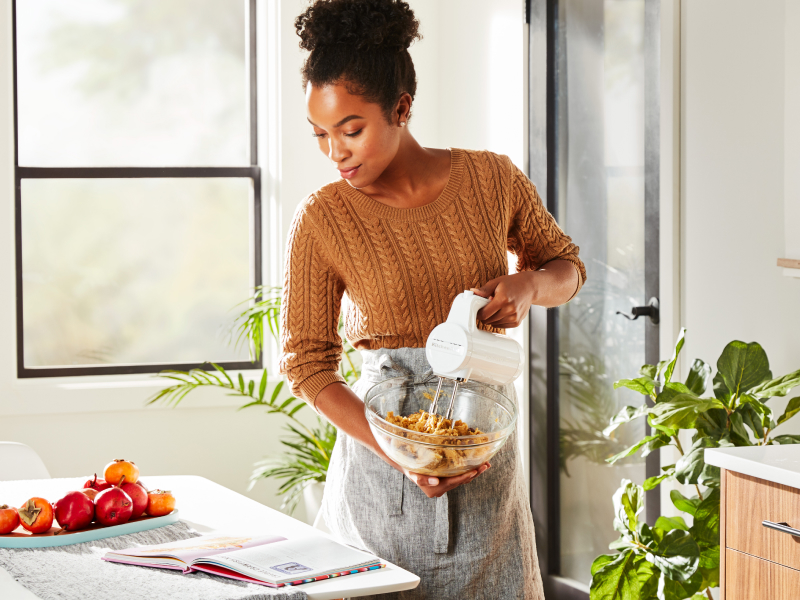 A woman in a modern kitchen reading a cookbook and holding a mixing bowl while using a Cordless KitchenAid® hand mixer to mix cookie dough.