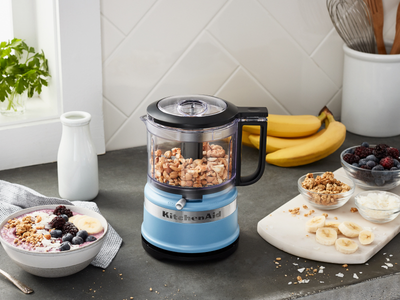 Yogurt and fruit next to a blue KitchenAid® food processor with nuts inside