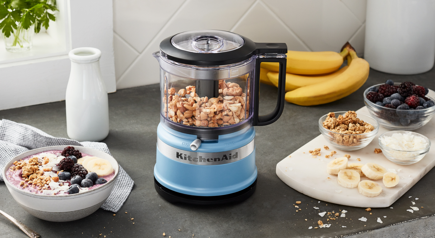 Yogurt and fruit next to a blue KitchenAid® food processor with nuts inside