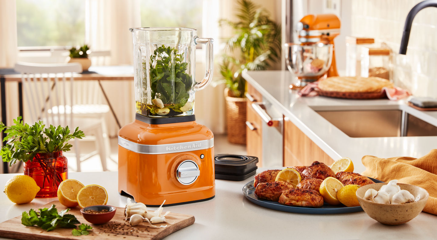 An orange KitchenAid® blender with sliced oranges and grilled chicken nearby.
