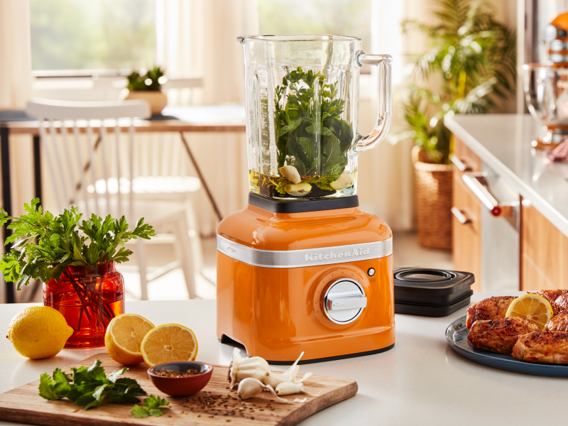 An orange KitchenAid® blender with sliced oranges and grilled chicken nearby.