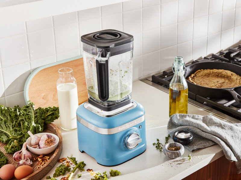 A KitchenAid® blender surrounded by various herbs and vegetable oil.