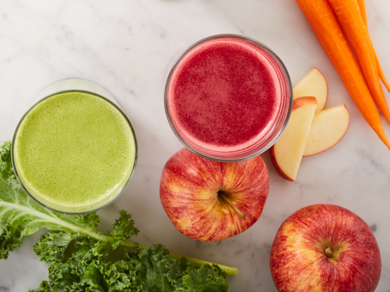 Green and red juices next to carrots, apples and lettuce on counter