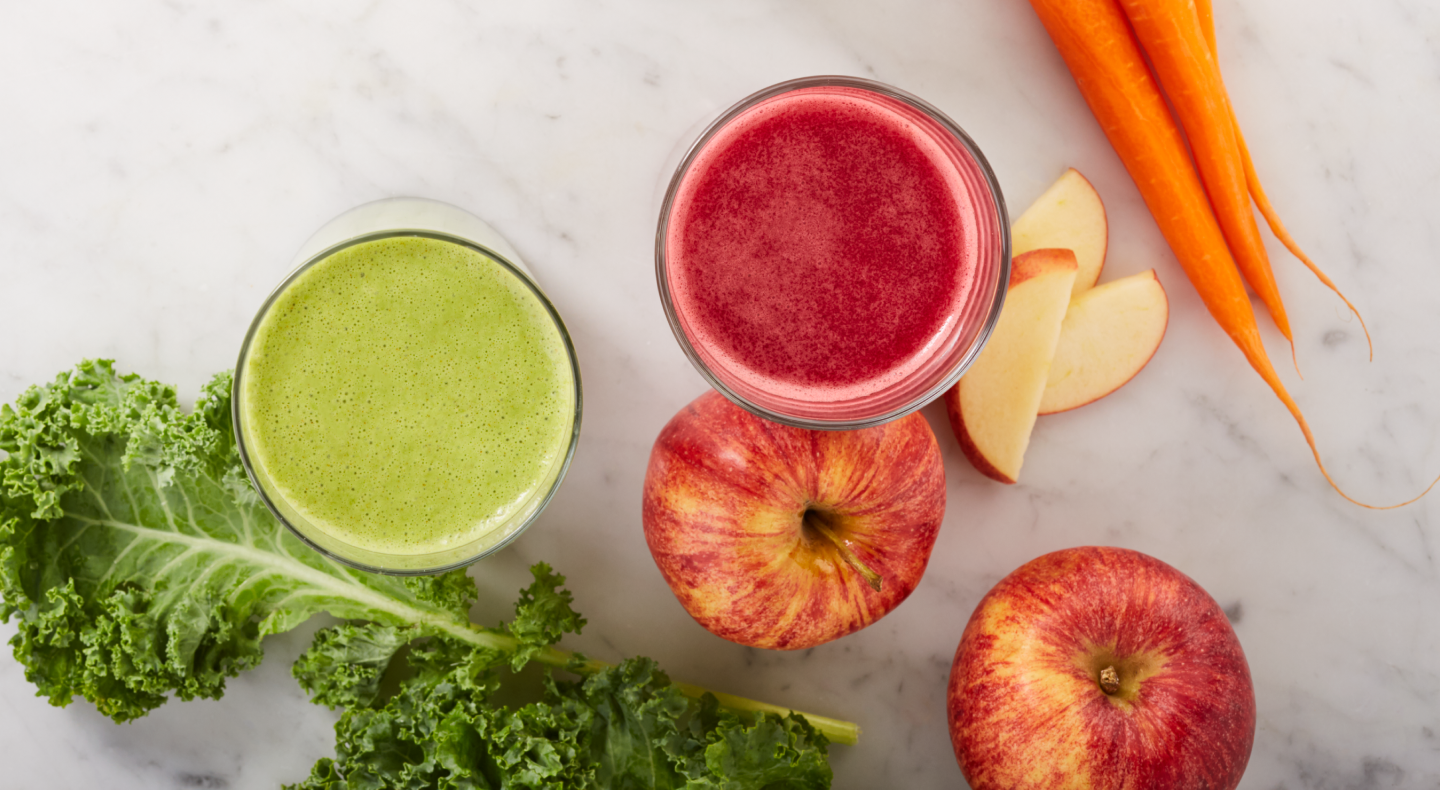 Green and red juices next to carrots, apples and lettuce on counter