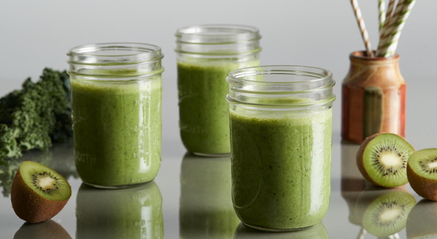 Mason jars of green juice on a countertop next to cut kiwis