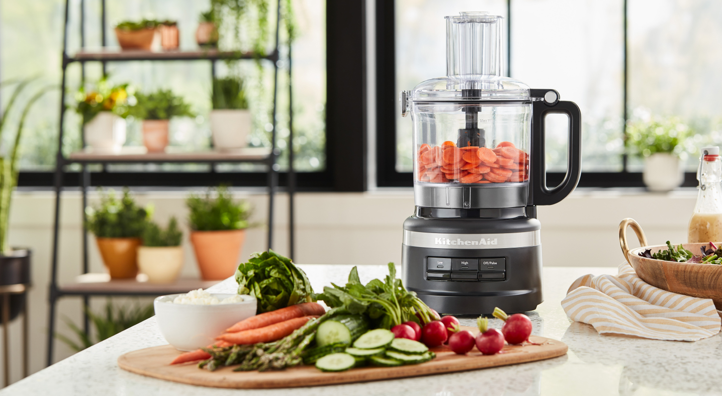 Cucumbers, carrots and radishes on cutting board next to food processor filled with sliced carrots