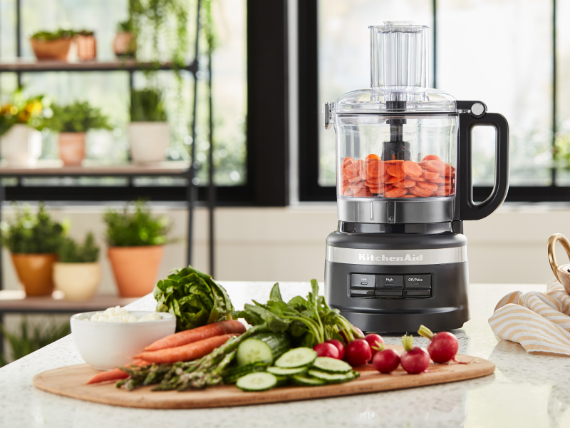 Cucumbers, carrots and radishes on cutting board next to food processor filled with sliced carrots