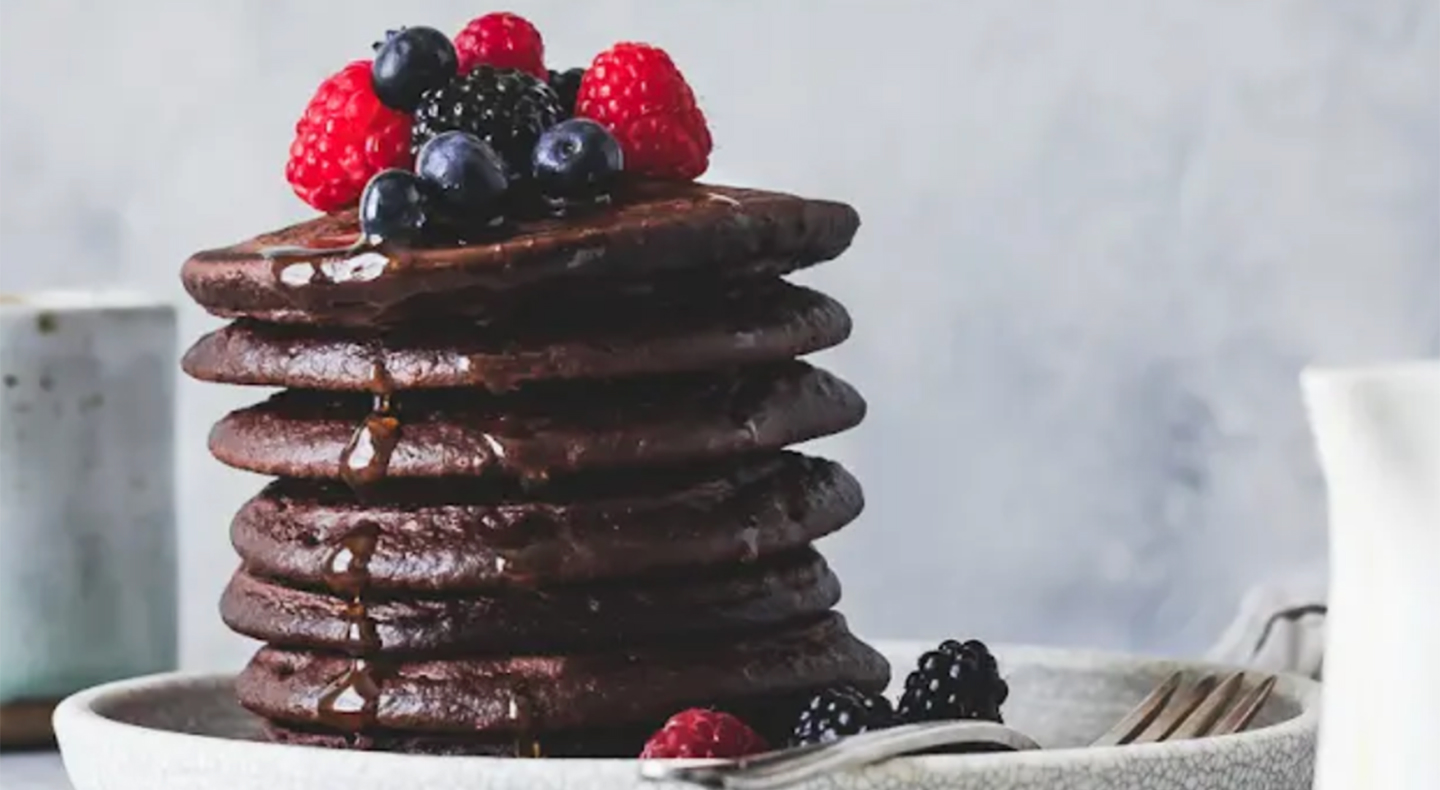 Stack of cocoa pancakes drizzled with syrup and topped with fresh fruit