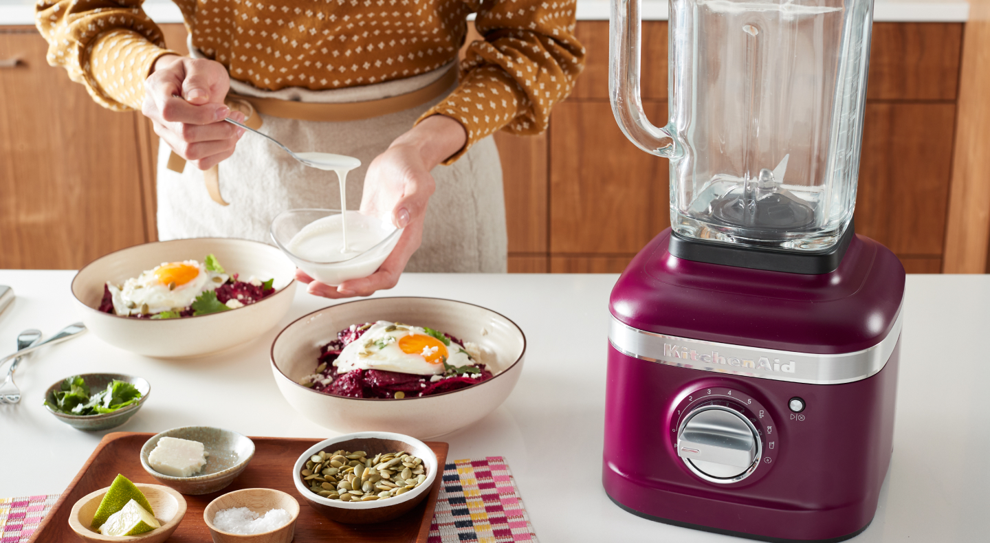 Person topping a harvest bowl with sauce next to a KitchenAid® blender
