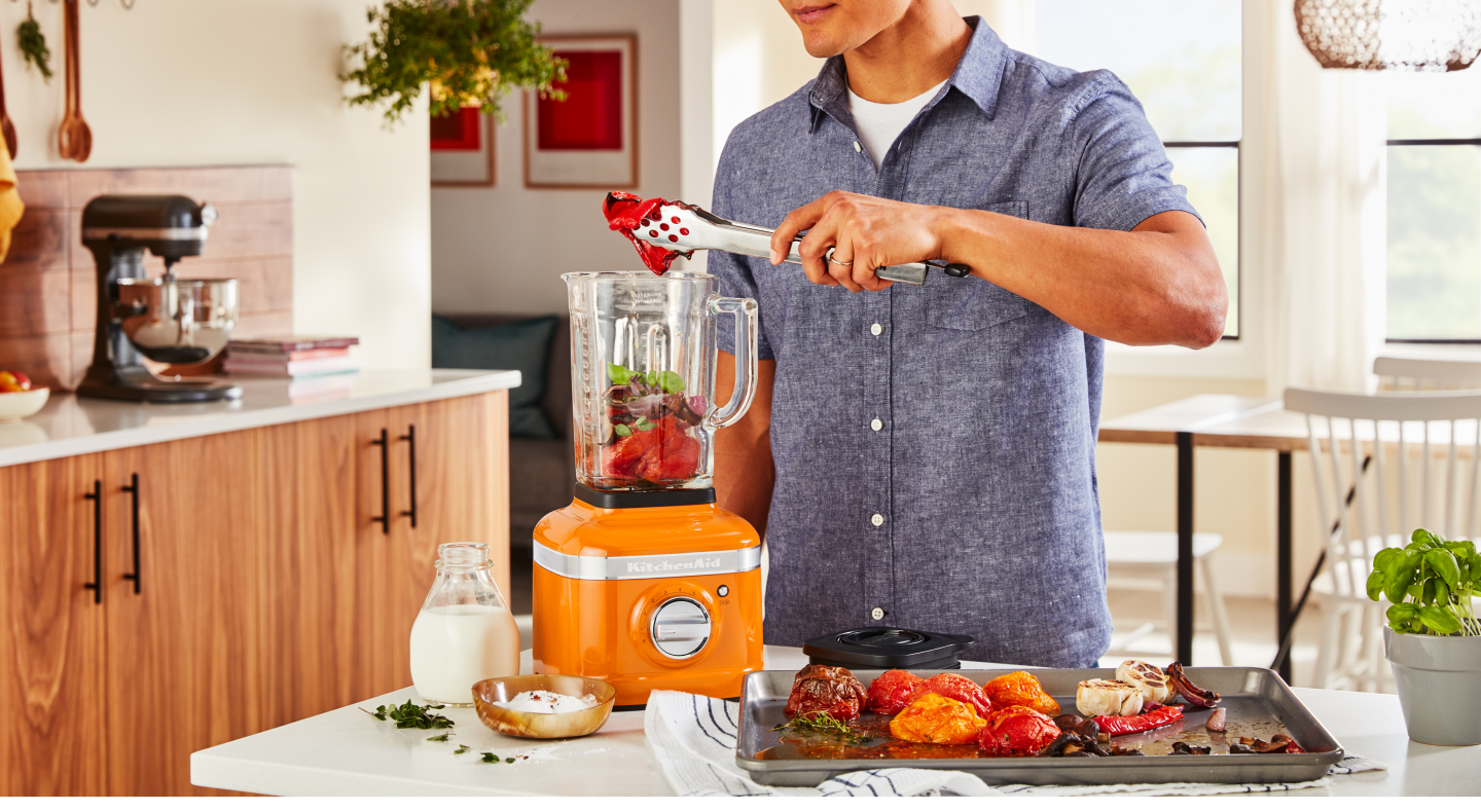 Person adding roasted vegetables into an orange KitchenAid® blender