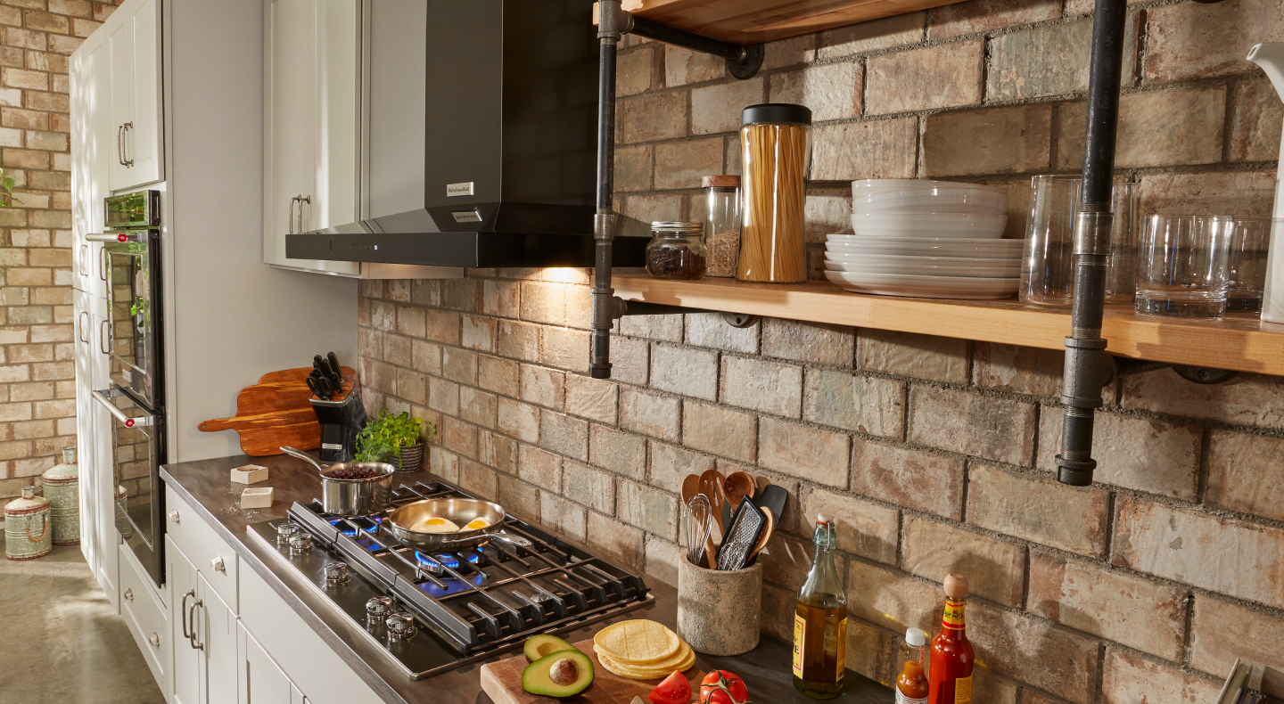 Black KitchenAid® canopy range hood over gas stovetop with brick backsplash