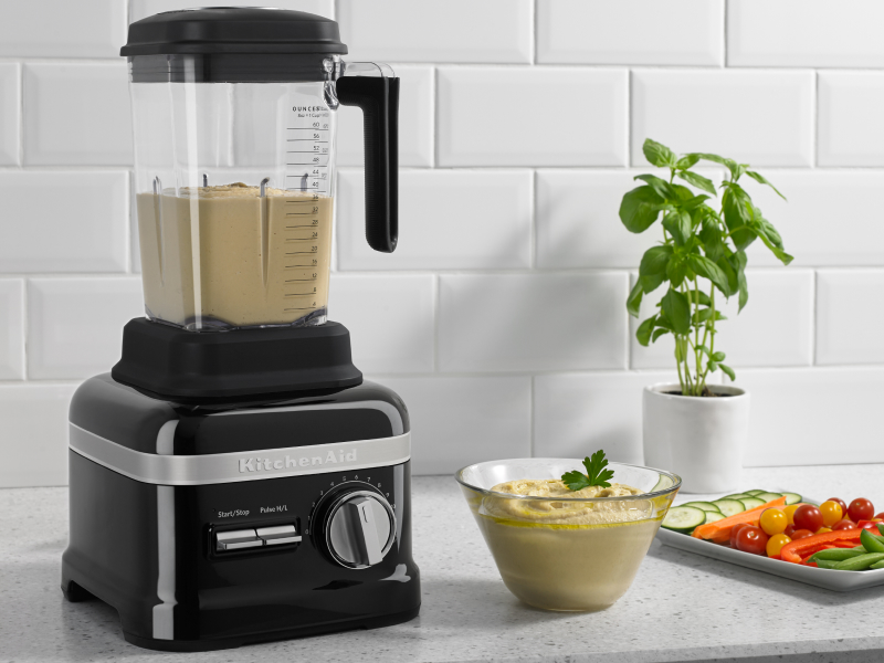 A KitchenAid® blender with hummus on a modern kitchen counter next to fresh vegetables.