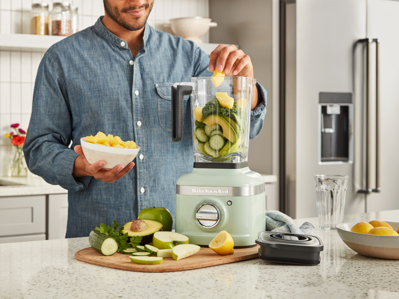 A man adding vegetables to a KitchenAid® blender in a modern kitchen.