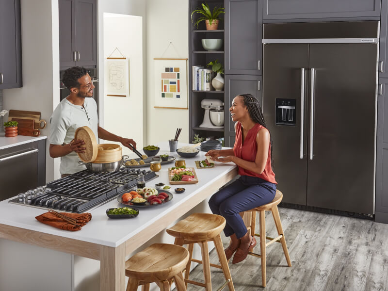 A man cooking for a woman on a downdraft gas cooktop set into a kitchen island
