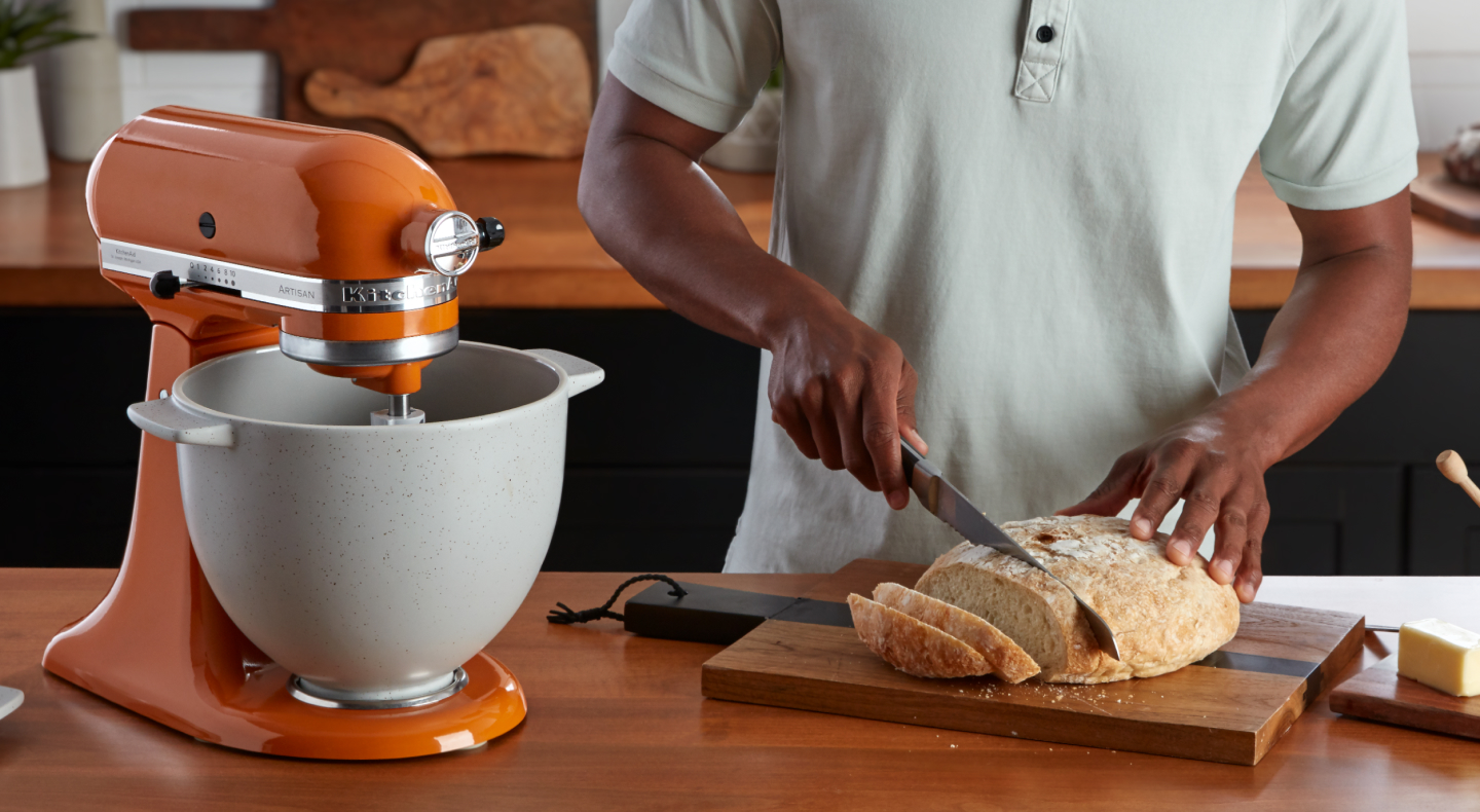 A person slicing bread next to an orange KitchenAid® stand mixer