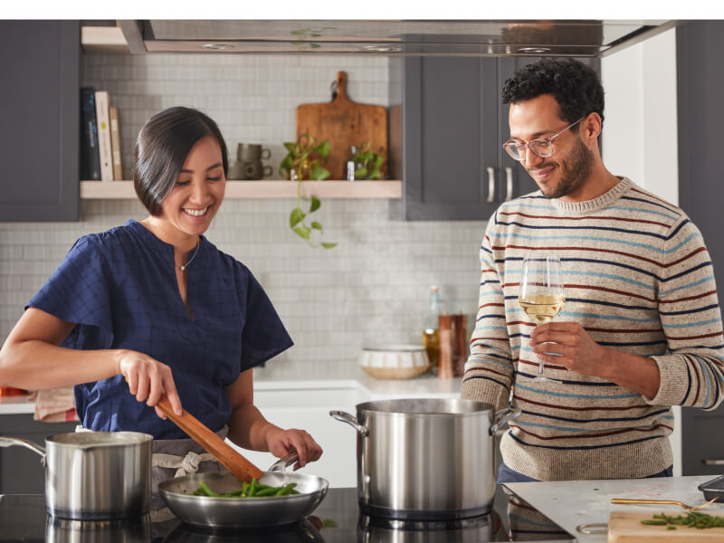 Two people cooking together using several stainless steel pans on an induction cooktop.