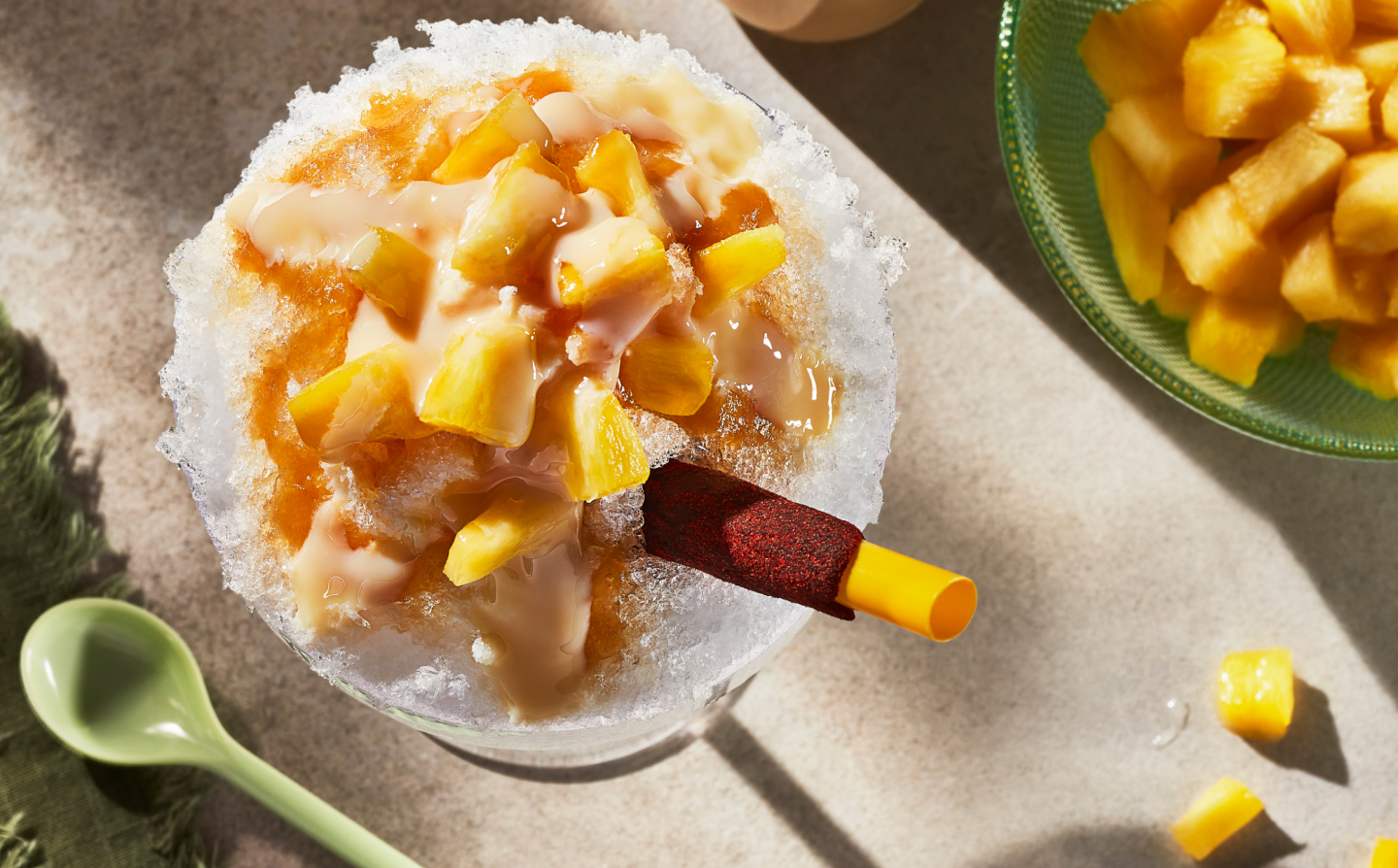 Shaved ice dessert topped with syrup and chopped fruit sitting next to green spoon