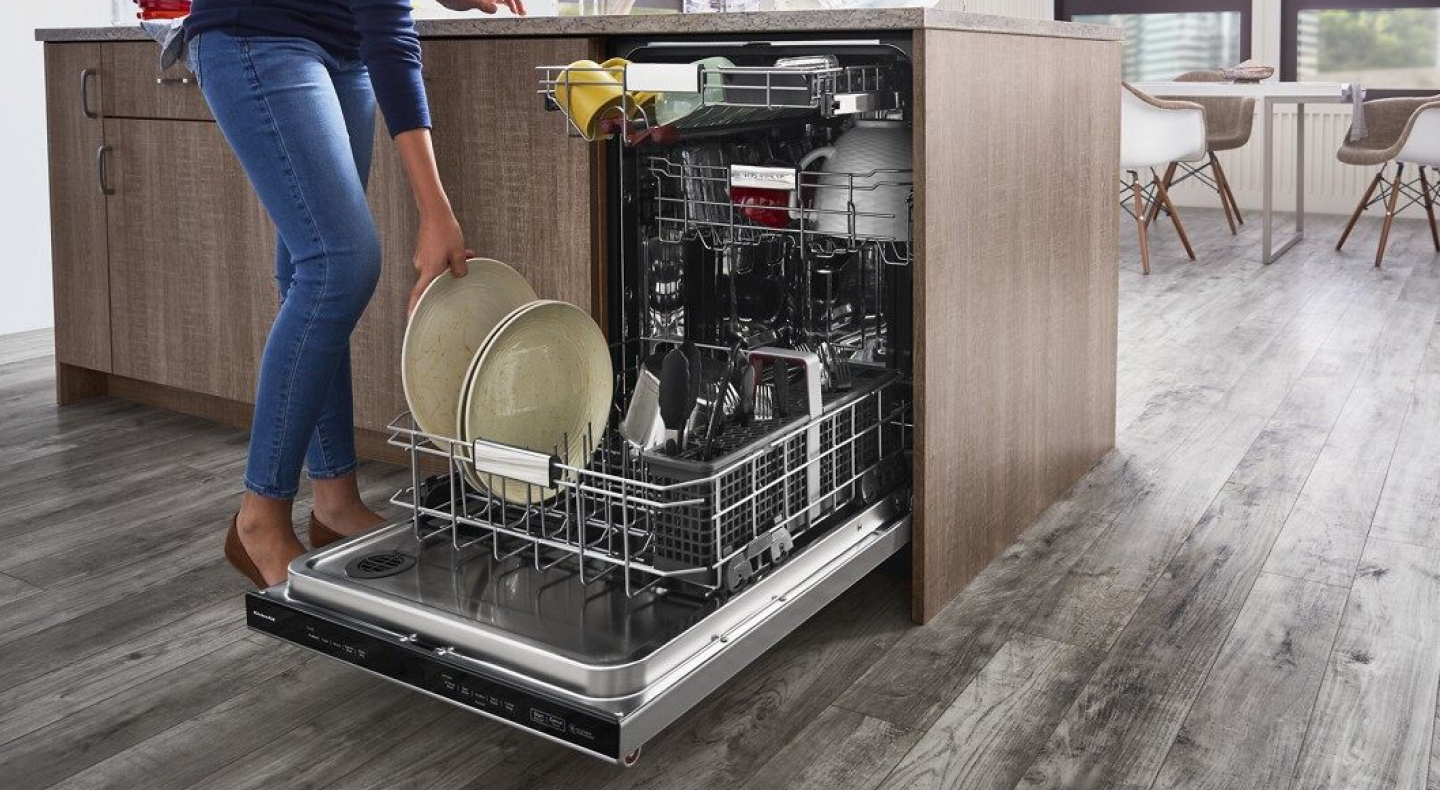 A closeup of a person loading plates onto the bottom rack of a stainless steel KitchenAid® dishwasher.