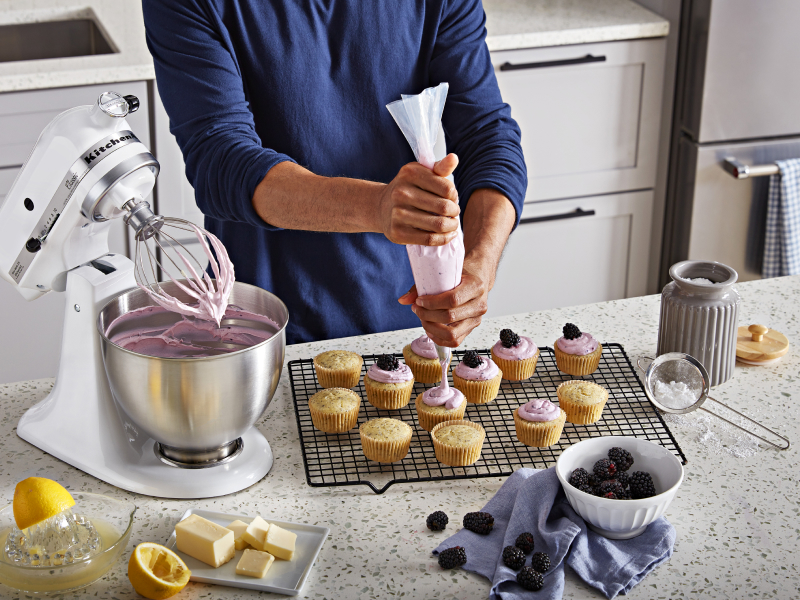 Person piping frosting onto cupcakes next to white stand mixer full of frosting