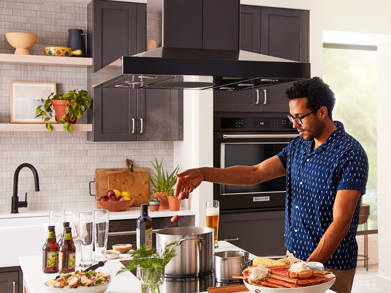 A person adding food to a pot on a KitchenAid® cooktop with a range hood above