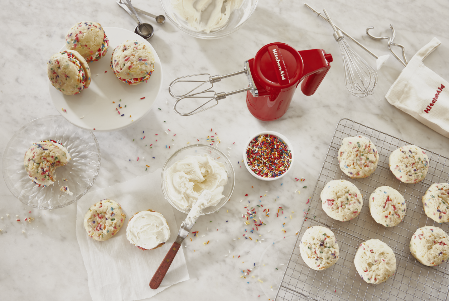 Red KitchenAid® hand mixer next to bowl of sprinkles, icing and bath of homemade cookie cream pies