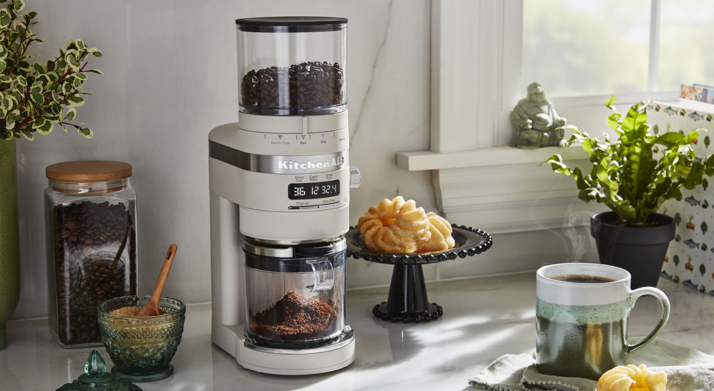 A KitchenAid® burr coffee grinder in a modern kitchen next to a pastry and cup of coffee.