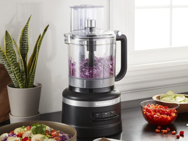 A black table that holds a food processor chopping onions as well as a fresh salad.