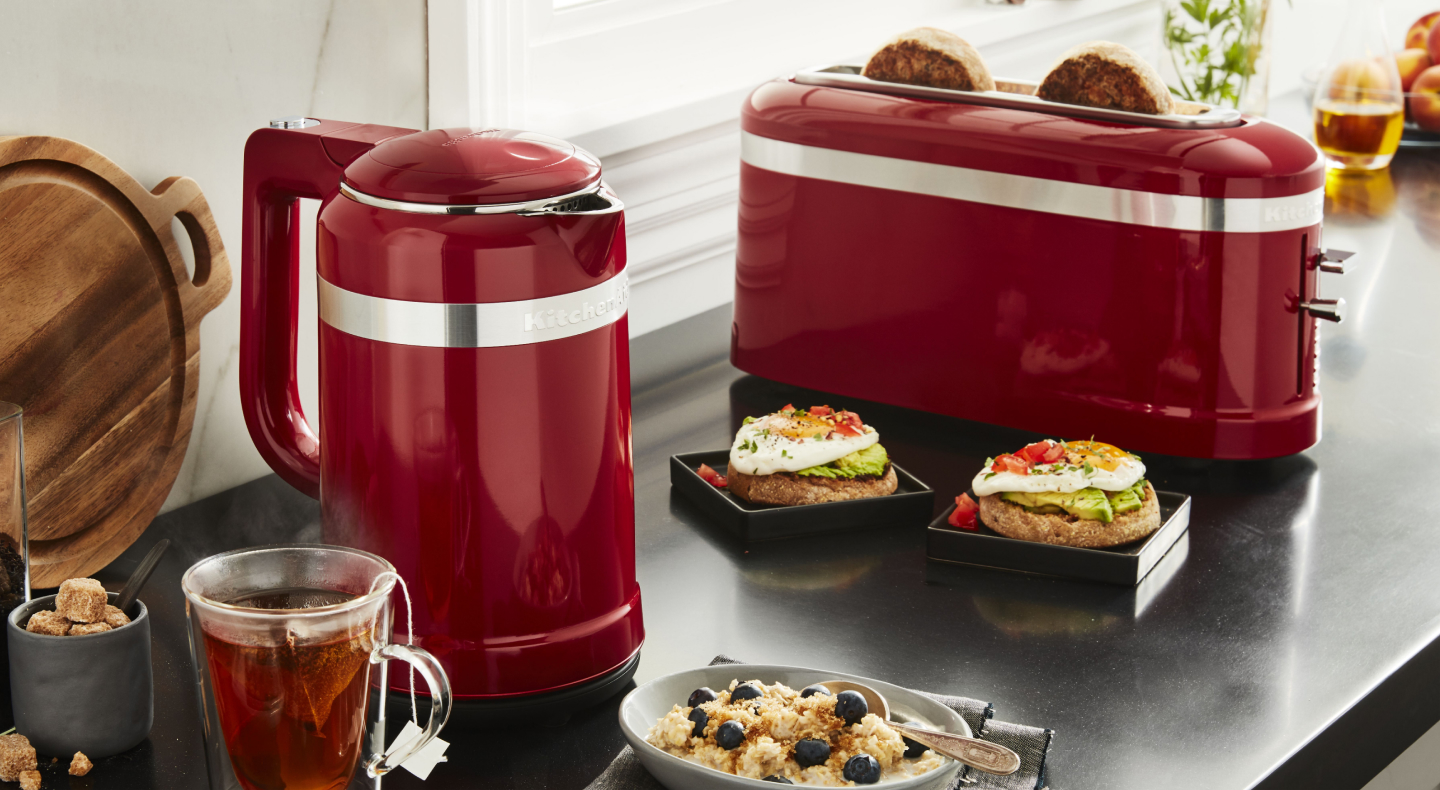 Slices of bread in a red KitchenAid® toaster next to open-faced sandwiches and cups of tea on a modern kitchen counter.