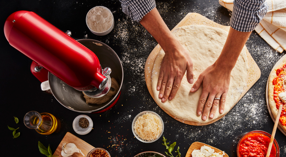 Two hands pushing down circle of dough on pizza peel next to red KitchenAid® Stand Mixer 