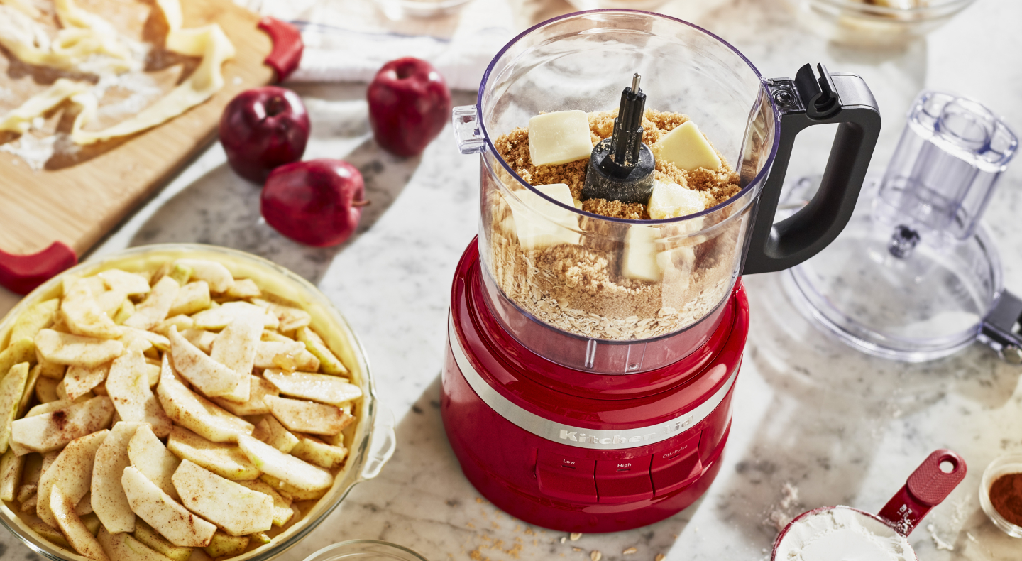 Crushed graham crackers and butter inside a KitchenAid® food processor next to an apple pie