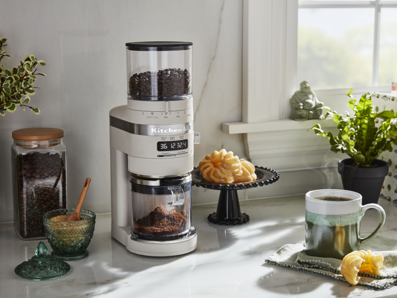 White KitchenAid® burr grinder next to a cup of coffee and pastries