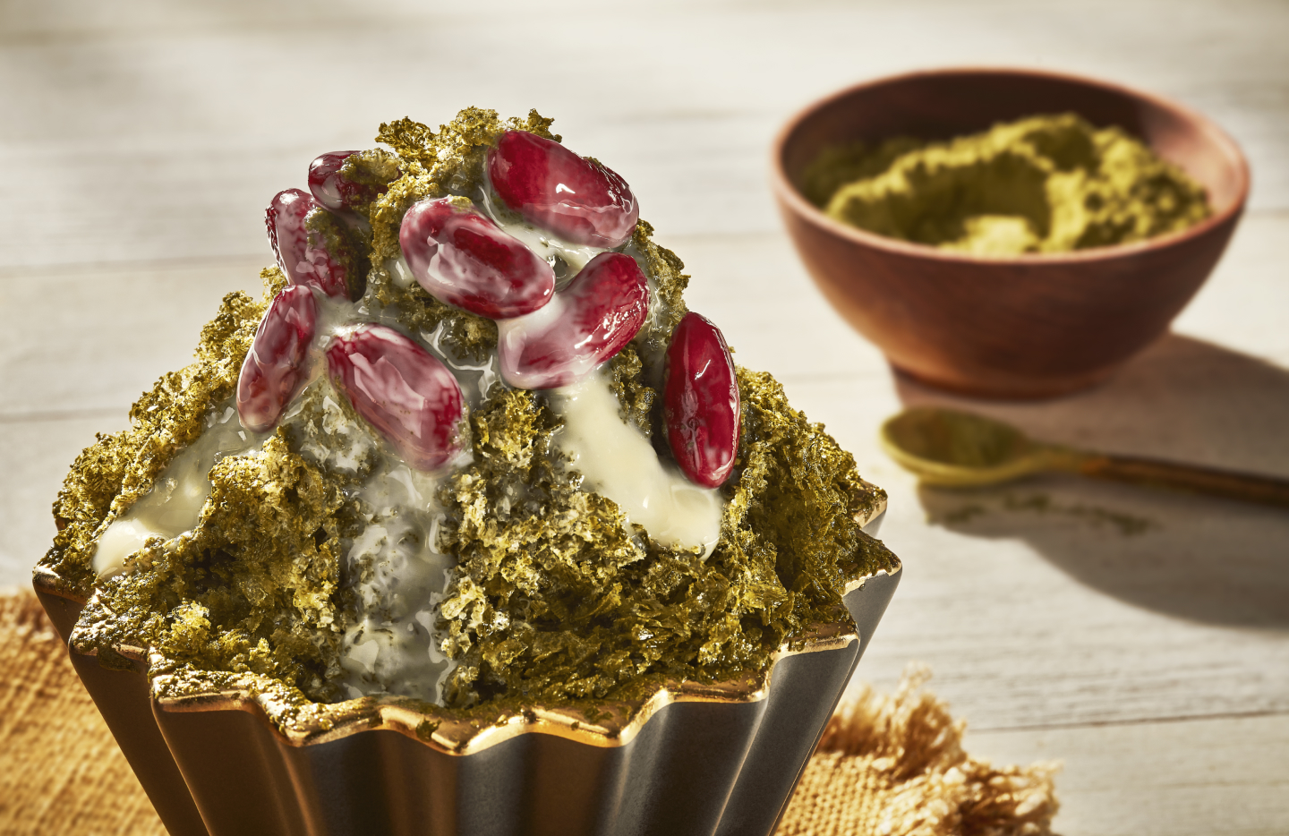 Shaved ice dessert with red beans and matcha powder beside bowl of match powder