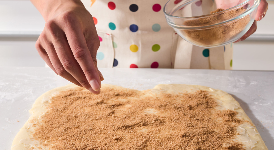 Person sprinkling filling onto cinnamon roll bread dough
